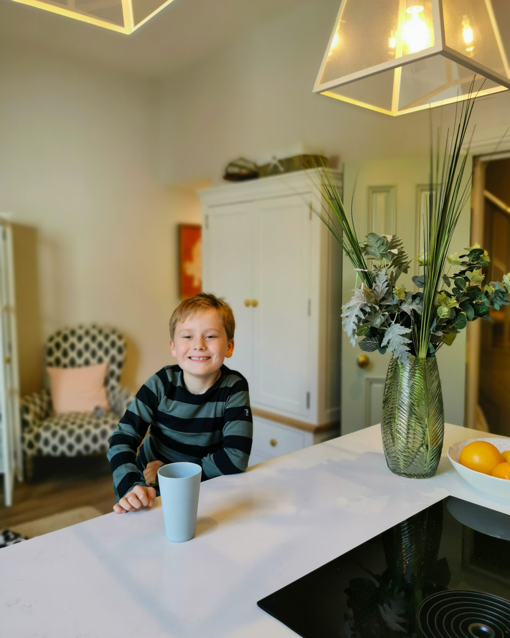white little boy wearing a striped top sitting on an island kitchen. He's got a cup in front of him and there is a vase with flowers and oranges in a bowl on the side