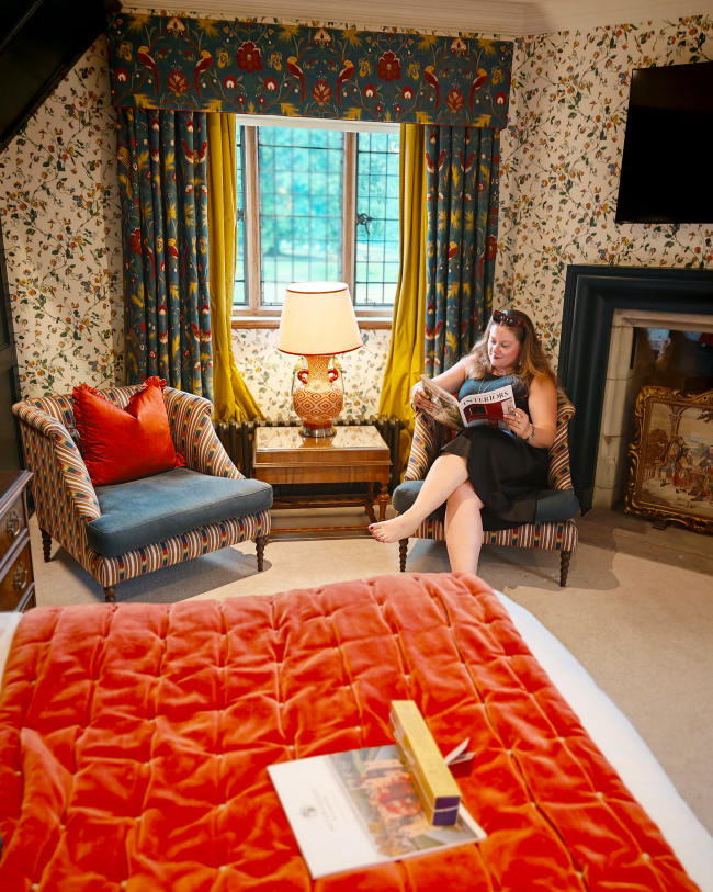 White woman in a black dress sat in a chair by the window in her room, reading a magazine. You can see the bed on the bottom of the photo, which is covered in an orange velvet throw