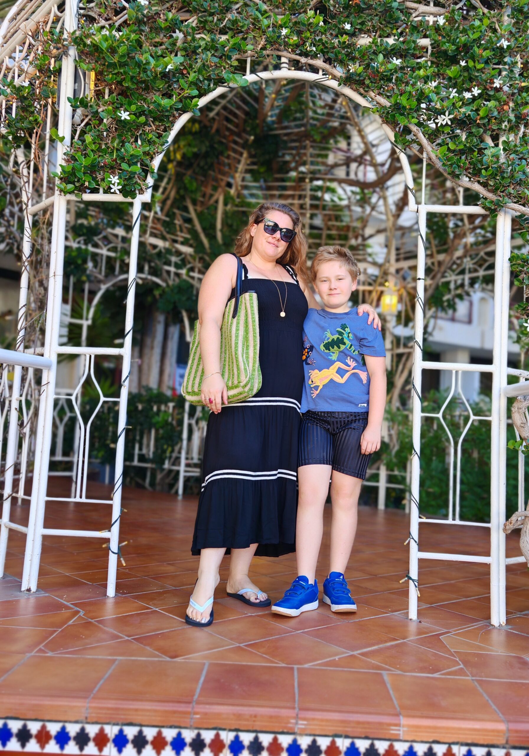 white woman on a long black dress with her son in a blue tee-shirt and black shorts, under an arched tree segment white woman on a long black dress with her son in a blue tee-shirt and black shorts, under an arched tree segment