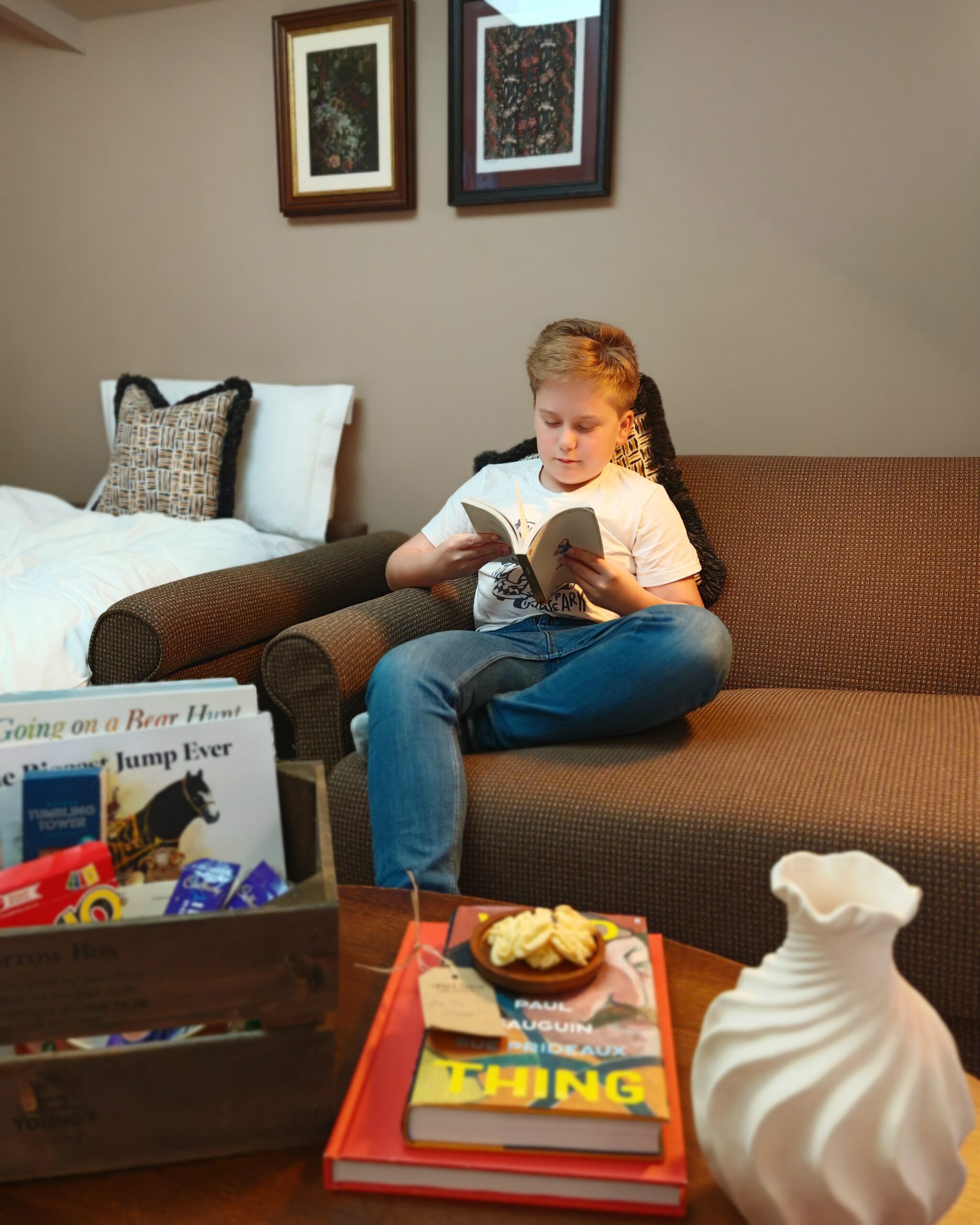 white little boy at on a sofa while reading a book. There are more books on the table in front of him and some games in a box white little boy at on a sofa while reading a book. There are more books on the table in front of him and some games in a box