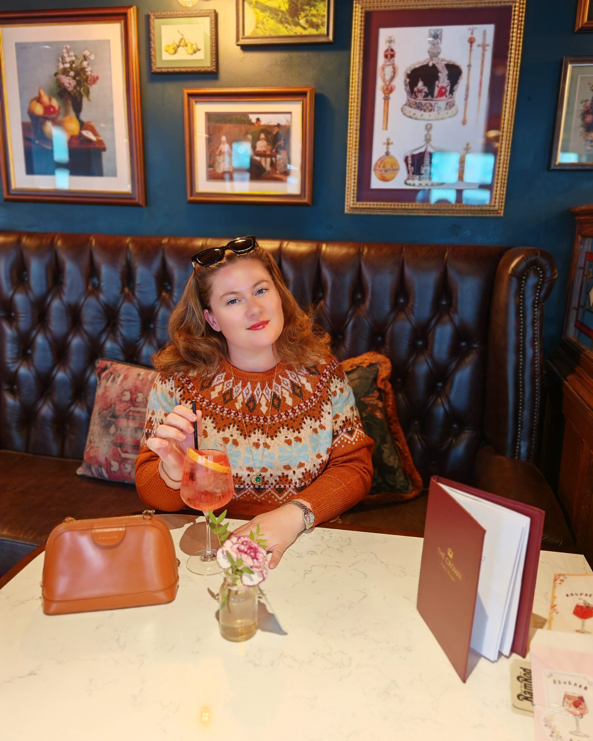 white woman wearing a brown fair isle crew jumper, sitting at a pub table with an aperol spritz cocktail white woman wearing a brown fair isle crew jumper, sitting at a pub table with an aperol spritz cocktail