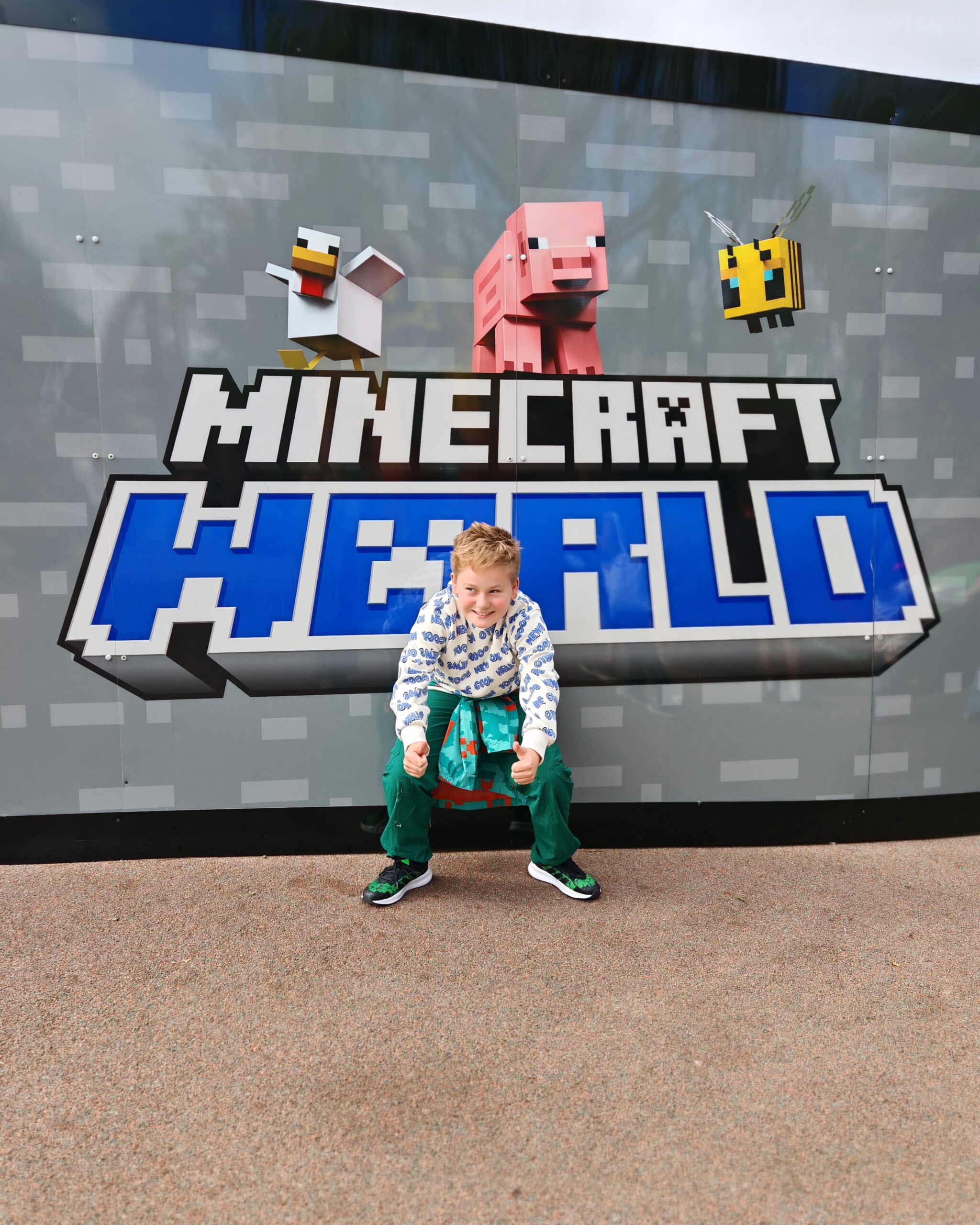 white little boy wearing a blue and white top posing in front of a minecraft sign announcing the new park coming to Chessington World of Adventures in Spring 2027 white little boy wearing a blue and white top posing in front of a minecraft sign announcing the new park coming to Chessington World of Adventures in Spring 2027
